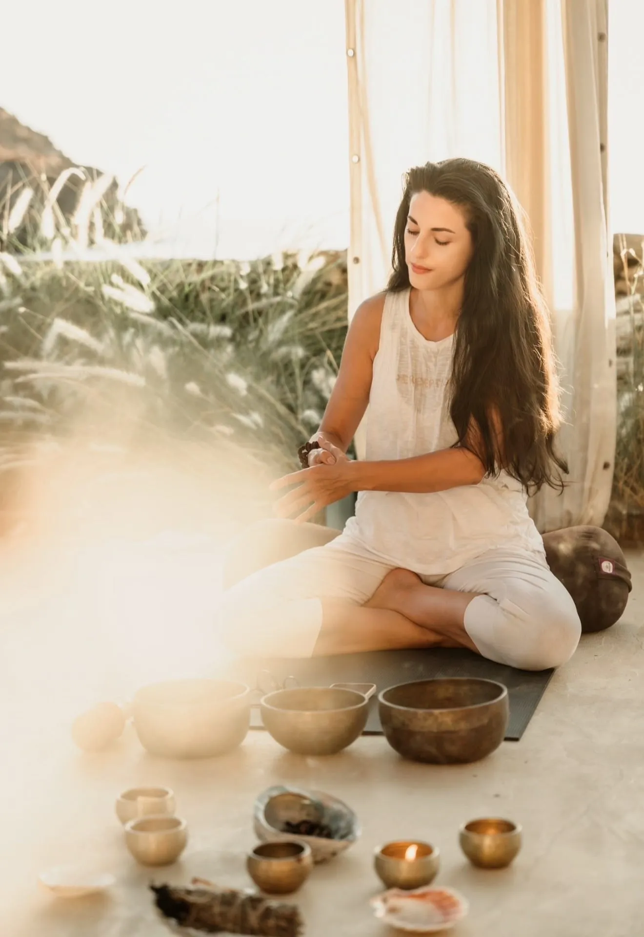 A practitioner seated in low evening sun beside a row of singing bowls, grasses behind