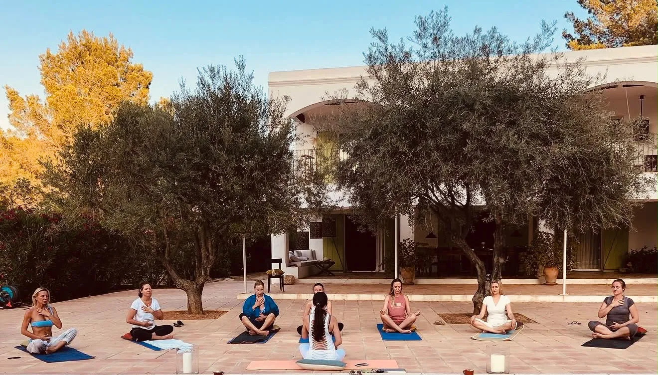 Dalia Lane guiding a seated group practice on a sunlit terrace beneath olive trees