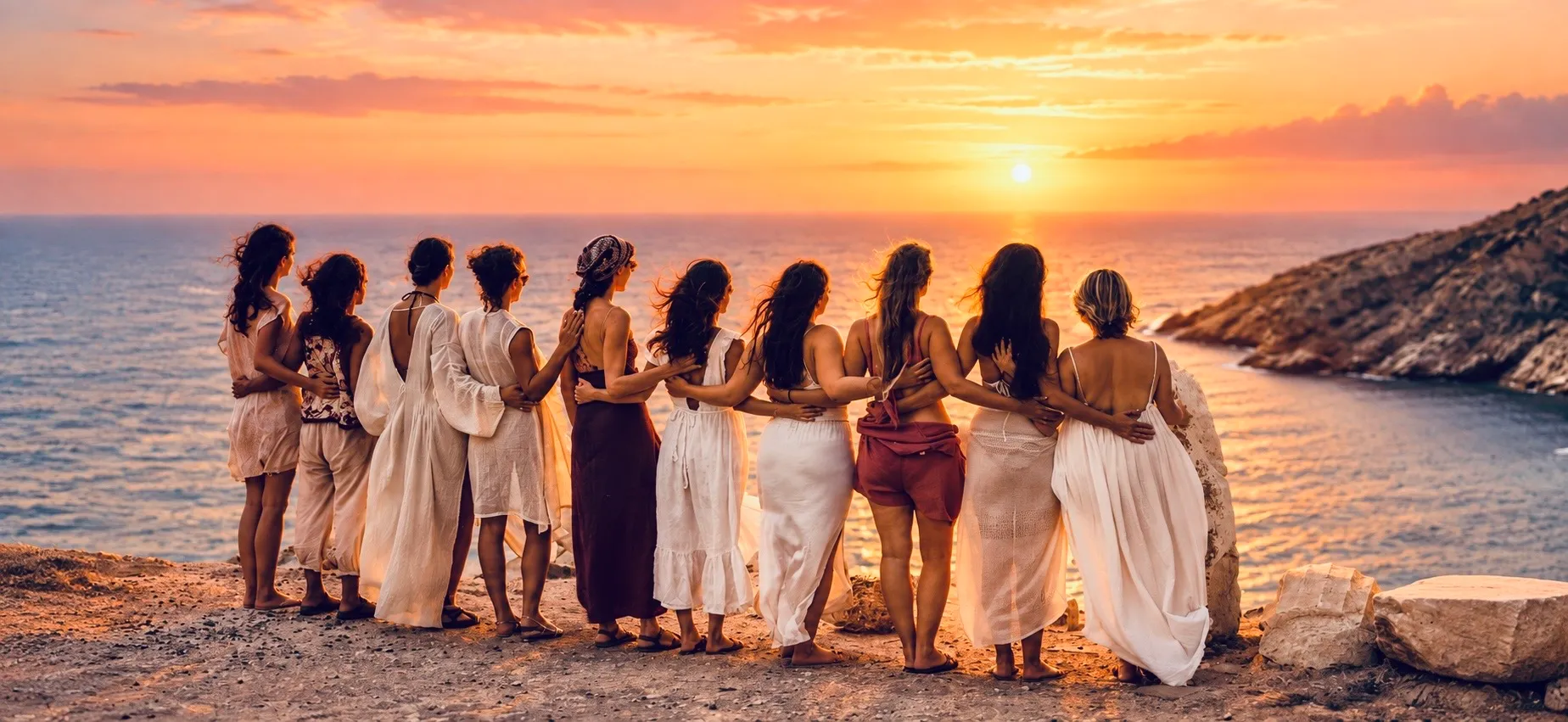 A circle of women with their arms around each other, looking out over the sea at sunset
